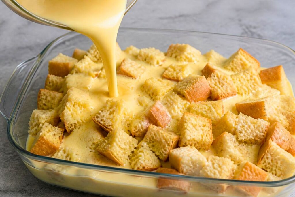 Creamy egg custard being poured evenly over cubed French bread in a baking dish for French toast casserole.
